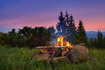 Bonfire in the tourist camp, water is heated in a metal mug. Beautiful summer evening, live fire warms tourists and helps to cook food