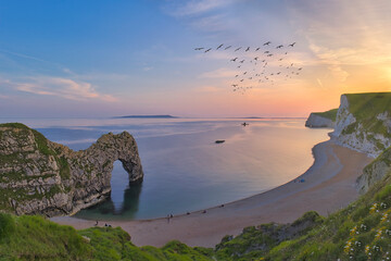 Durdle Door Beach with a flock of birds in the purple and magenta sunset sky and a silhouette of a...