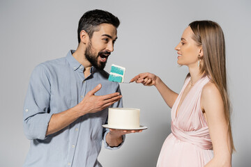 Smiling pregnant woman in pink dress holding blue cake near husband with open mouth during gender party celebration on grey background, expecting parents concept, it`s a boy