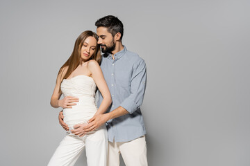 Trendy and bearded man hugging fair haired and pregnant woman while touching belly while standing together on grey background, expecting parents concept, baby bump, husband and wife - Powered by Adobe