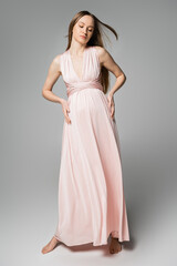 Full length of barefoot long haired and pregnant woman in pink dress touching belly while standing...
