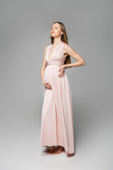 Full length of relaxed and fair haired pregnant woman in pink dress touching belly while standing on grey background, elegant and stylish pregnancy attire, sensuality, mother-to-be