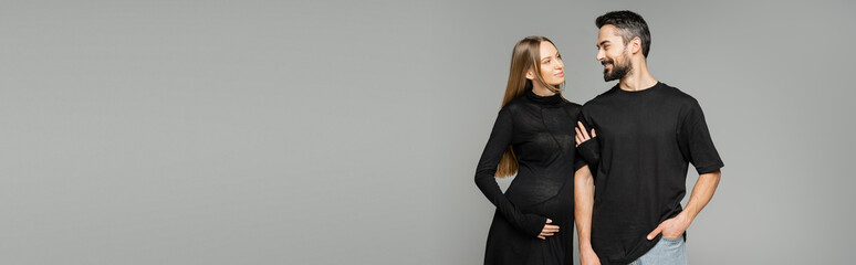Stylish and pregnant woman in black dress touching arm of smiling bearded husband and standing together isolated on grey, new beginnings and parenting concept, banner