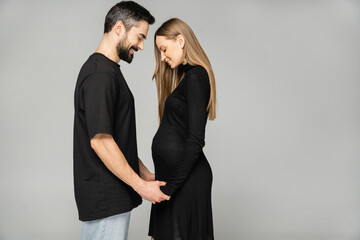 Side view of positive pregnant and stylish woman in black dress holding hands of bearded man while standing isolated on grey, new beginnings and anticipation concept, husband and wife