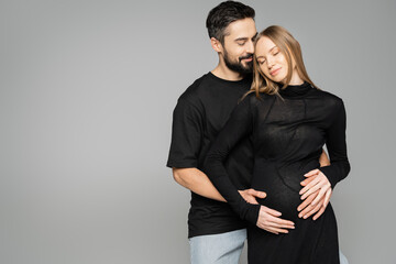 Positive bearded husband in black t-shirt hugging stylish pregnant wife in dress and closing eyes and standing isolated on grey, new beginnings and anticipation concept