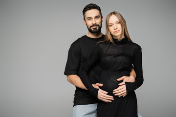 Bearded and smiling man in black t-shirt hugging stylish pregnant wife in dress and looking at camera while standing isolated on grey, new beginnings and parenthood concept