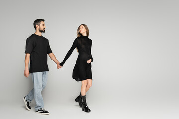 Full length of trendy pregnant woman in dress walking and holding hand of bearded husband in t-shirt and jeans on grey background, new beginnings and anticipation concept, expecting parents