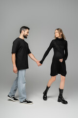 Full length of stylish and pregnant woman in black dress holding hand of smiling husband and walking together on grey background, new beginnings and anticipation concept