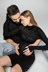 Trendy and pregnant fair haired woman in black dress hugging bearded husband while sitting together on grey background, new beginnings and parenting concept