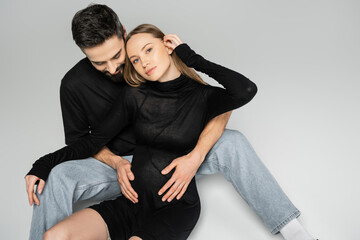 Husband in black t-shirt and jeans touching belly of fashionable pregnant wife in dress looking at camera while sitting on grey background, new beginnings and parenting concept
