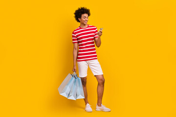 Full length photo of funny pretty man wear striped t-shirt holding shopper texting iphone gadget isolated yellow color background