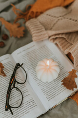 Fototapeta na wymiar White burning candle shape of pumpkin on open book, eyeglasses, cozy sweater and autumn leaves on bed
