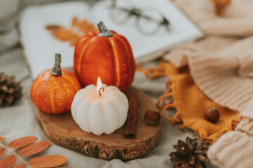 Fototapeta na wymiar Cozy autumn decor - burning candle shape of pumpkin and orange decor pumpkins on wooden board on bed with open book, warm sweater, autumn leaves