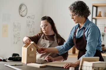 Girl with down syndrome making birdhouse together with carpenter in workshop