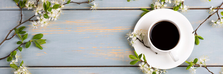 Cup of fresh black coffee and cherry blossom branches on blue wooden background. Coffee break. Spring flowers. Flat lay, top view, banner