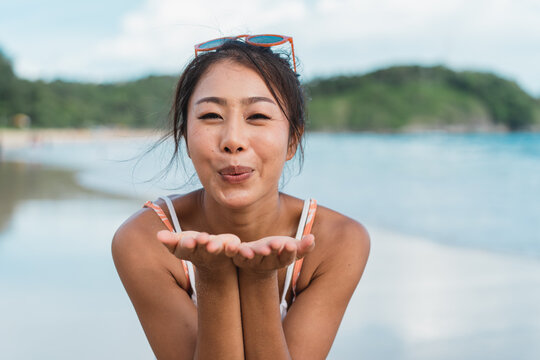 Smiling beautiful Asian woman blowing a kiss in bikini on the beach and sea in background. Pretty young playful female traveler in swimwear having fun in summer time.