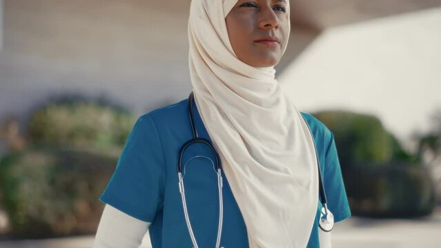 A young Muslim woman, a doctor in a hijab in uniform with a stethoscope, is standing on the street outside the hospital. Medicine and health care.