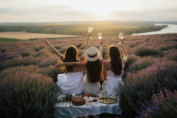 Girlfriends having picnic in the lavender field at sunset. Group of young women sitting on lavender field on summer day. Girlfriends drinking wine on outdoor party.	
