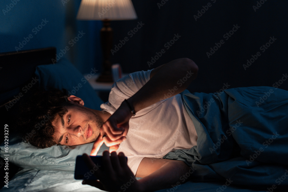 Wall mural Portrait of young sleepy man lying in bed using smartphone at late night, no sleep. Insomnia, sleep disorder concept. Dependency on a cellphone. Loneliness crisis - Wall murals