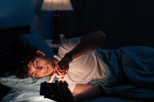Portrait of young sleepy man lying in bed using smartphone at late night, no sleep. Insomnia, sleep disorder concept. Dependency on a cellphone. Loneliness crisis