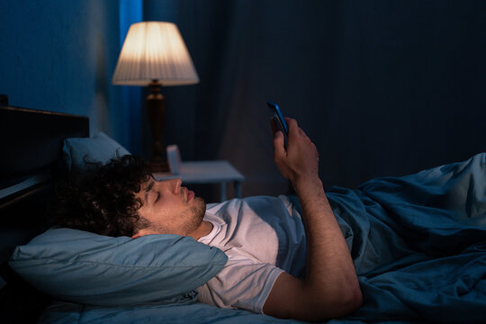 Dark night young man looking at mobile phone screen texting late awake in bed insomnia. Serious guy depressed addicted to social media.