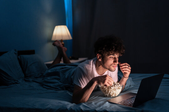 Happy muslim student eating popcorn while lying on bed watching movie on laptop at night at home