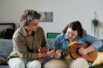 Teacher teaching girl with down syndrome to play guitar while they sitting on sofa in the room