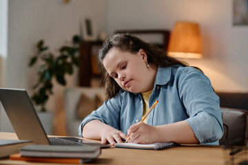 Naklejka na ściany i meble Girl with down syndrome making notes during online lesson while sitting at desk in front of laptop