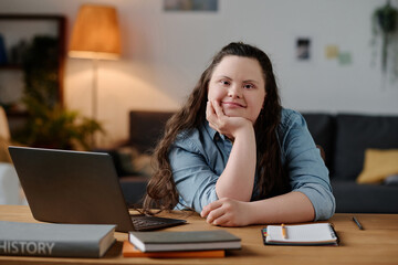 Fototapeta Portrait of girl with down syndrome looking at camera while sitting at desk with laptop in the living room obraz