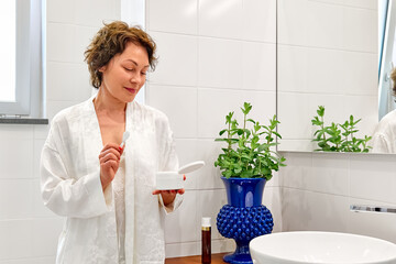 Middle-aged woman in white silk bathrobe in bathroom in front of mirror applying hydrating anti-aging cosmetics on skin of her hands and smiling.