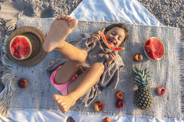 Little girl eats fruit lying on a blanket on the beach.