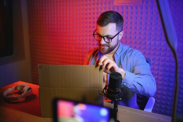 Fototapeta na wymiar Panoramic view of young adult vlogger creating internet content, holding carton box in hands, unpacking received pack and recording media online video on broadcast equipment