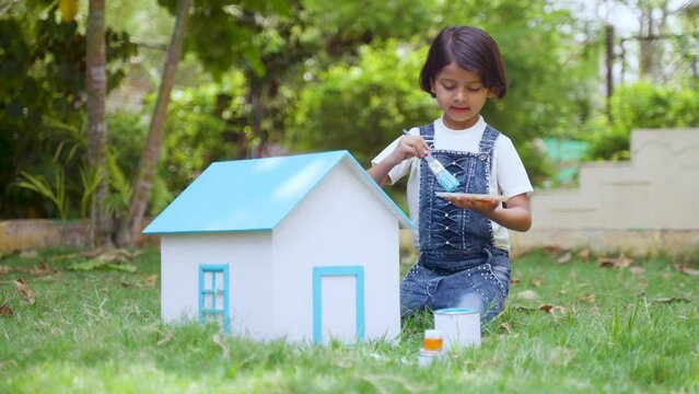 Happy indian girl kid painting cardboard toy house using brush at park - concept of childhood creativity, playful and inspiration