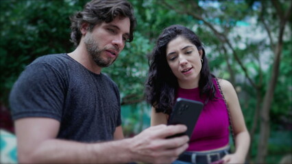 Young man showing online content to woman on cellphone device, both standing outside at city park. Two people looking at smartphone screen comenting on the media they are watching