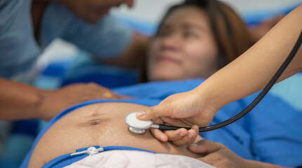 male obstetrician doctor in white coat gown use stethoscope to check womb stomach in delivery room. husband encouraging soothe wife from womb pregnant abdomen in Labour room