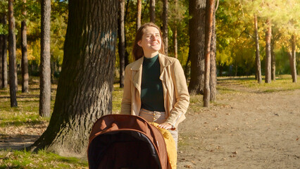 Portrait of happy smiling woman with long hair walking in park with her baby sleeping in stroller. Parenting, relaxing outdoors, beautiful mother