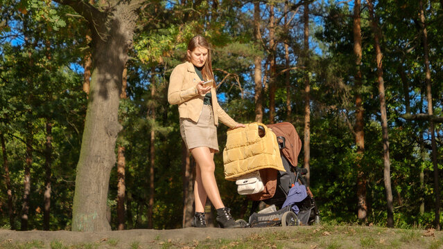 Young woman using smartphone while walking with baby stroller in park. Parenting, relaxing outdoors, beautiful mother