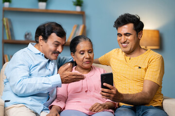 Happy Indian adult son showing mobile phone to senior parents while sitting on sofa at home - concept of matrimony app, technology and family bonding