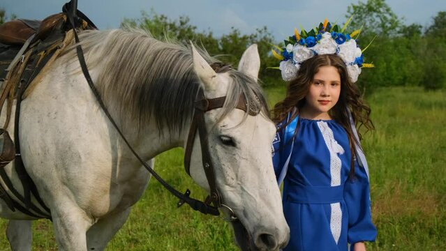 A beautiful young girl leads a white stallion. The girl is dressed in a national Ukrainian dress. Close-up.