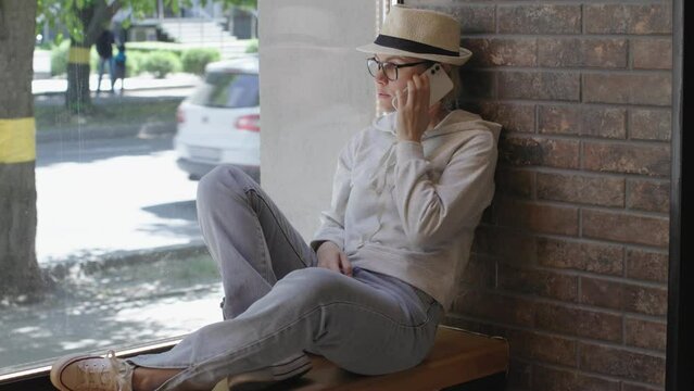 happy lifestyle city town cafe panoramic window female sitting windowsill wear blue jeans white hat and glasses talking on phone looking through window cars driving summer day. tourist woman freelance