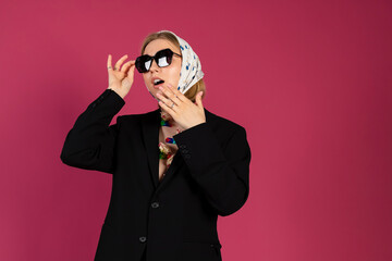 Portrait of a Beautiful tender young blonde hair girl is wearing a stylish black blazer and creative flowers attached to her chest and babushka head scarf. Isolated on pink background.