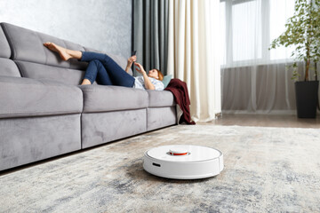 The girl lies on the sofa in the living room and controls a smart robot vacuum cleaner using a smartphone.