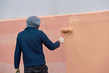 Painting wall with paint roller outdoors, removing graffiti from building. Worker manually painting pink wall, renovation facade works. Industrial painter at work, vandalism concept.