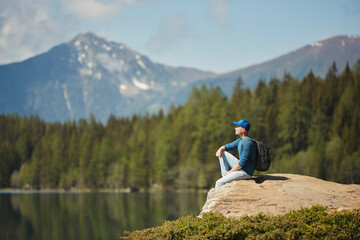 Man with backpack sitting on rock and enjoying scenic view of mountain lake. Young solo traveler in the Alps..