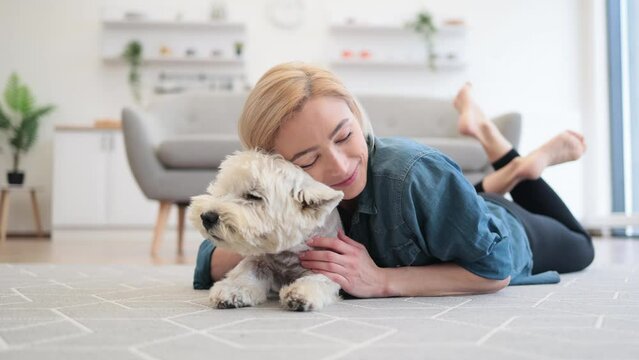 Pretty smiling woman in casual wear snuggling to cute pet's head while testing comfort of wooden floor with carpet on it. Joyful healthy lady appreciating affection and support of her canine friend.