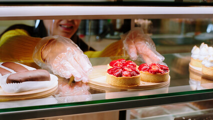 In the coffee shop saleswoman with protective mask arrange the deserts from the showcase fridge very carefully