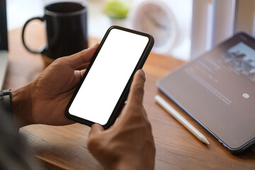 Creative man hand holding smartphone with white empty screen at wooden working desk.