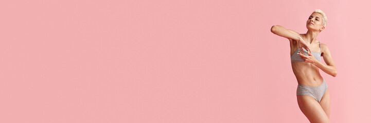 Naklejka premium Portrait of tender, young blonde woman with slim body posing i cotton grey underwear against pink studio background