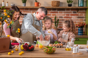 Obraz na płótnie Canvas In the morning happy young family cooking their favourite deserts the dad charismatic man mixed the dough while other family members helping him at the kitchen island