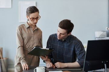 Portrait of two young professionals looking at clipboard while reviewing IT project in office, copy space